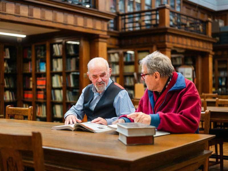 To ældre mænd sidder sammen og taler i et bibliotek.