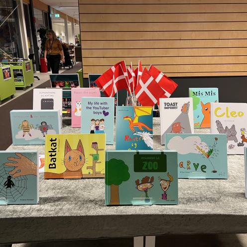 Bøger i forskellige farver og formater med børnenes tegnede forsider er udstillet på et bord med flag og dug.