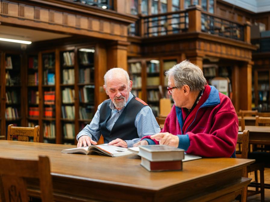 To ældre mænd sidder sammen og taler i et bibliotek.